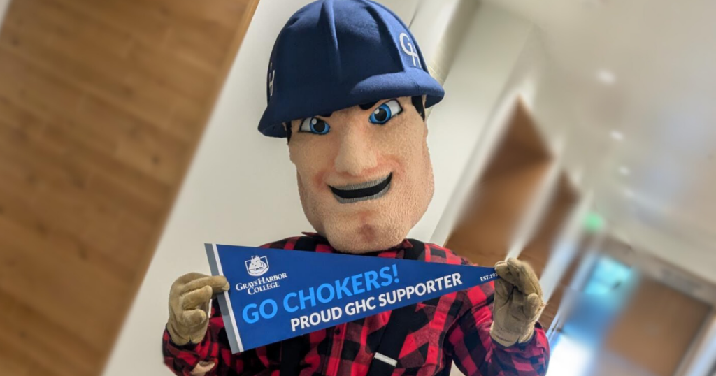 The Grays Harbor College Charlie Choker mascot, dressed as a lumberjack with a blue hard hat and plaid red-and-black shirt, is holding a blue pennant that reads "GO CHOKERS! PROUD GHC SUPPORTER" with the Grays Harbor College logo. The mascot is smiling and posing in a hallway with wood and white accents.