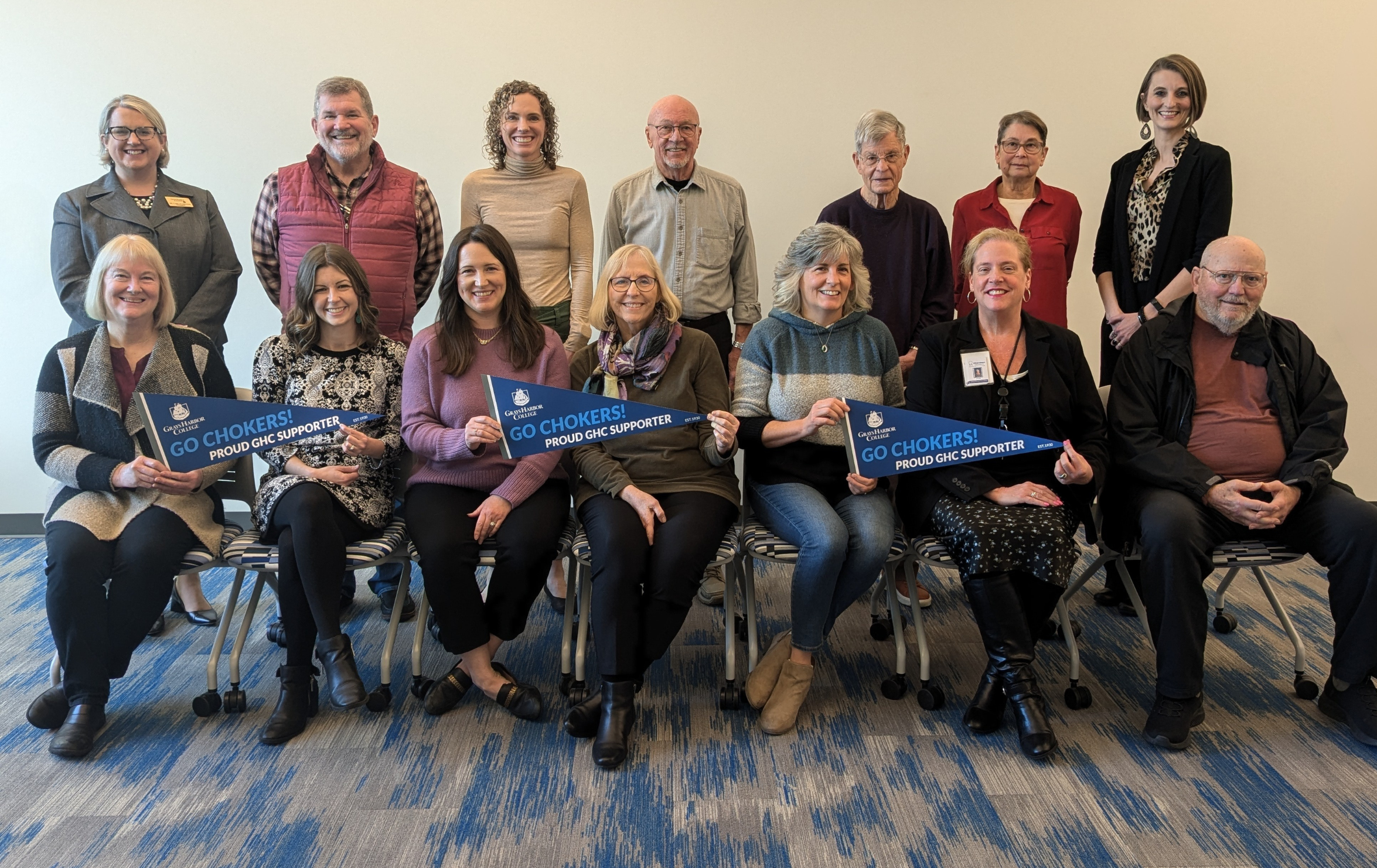 A group photo of the Grays Harbor College Foundation Board of Directors. The image features thirteen individuals posed in two rows against a light-colored wall. The back row includes six standing individuals, while the front row has seven seated individuals. Several people in the front row are holding blue pennants that read, "Go Chokers! Proud GHC Supporter." The group is diverse in appearance, dressed in business-casual attire, and is seated on a patterned blue carpeted floor. The setting appears professional and welcoming.