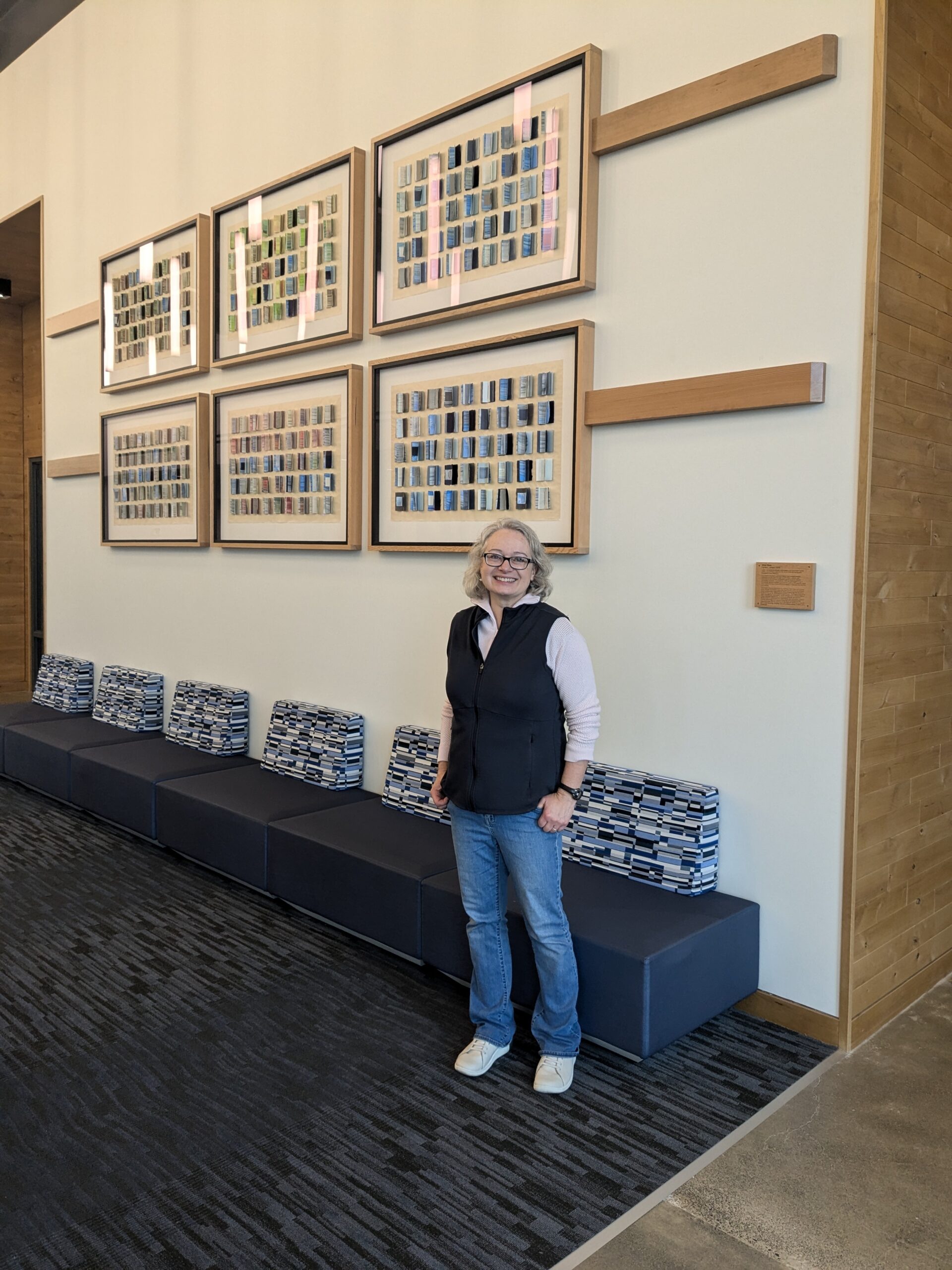 Artist Carrie Larson stands in front of her commissioned art piece, Vital Flow, which features a collection of framed, multicolored rectangular elements arranged in a grid pattern on a light-colored wall.