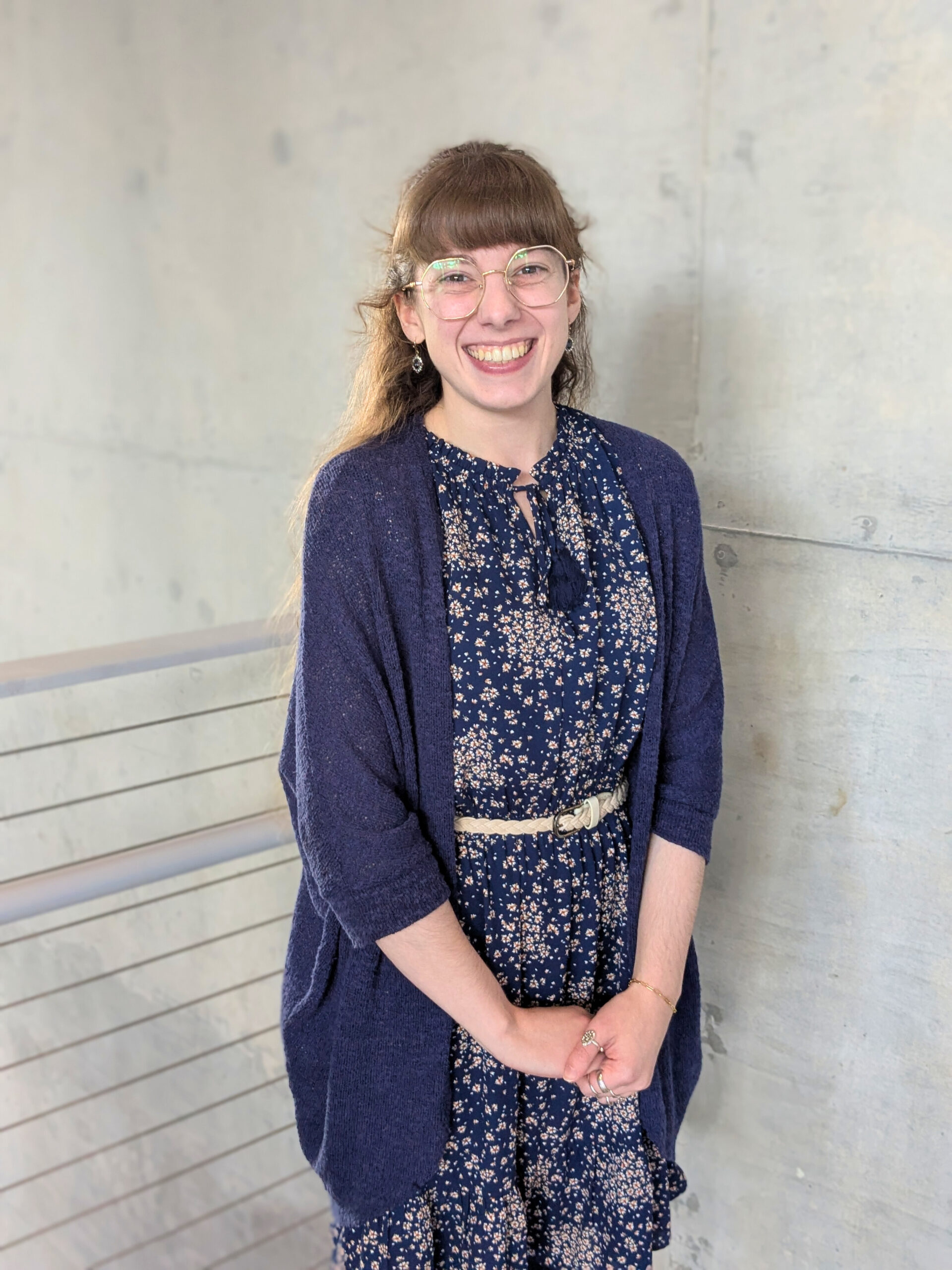 Program Manager, Bryn Cavin, standing against a gray wall, wearing a blue outfit, smiling warmly.