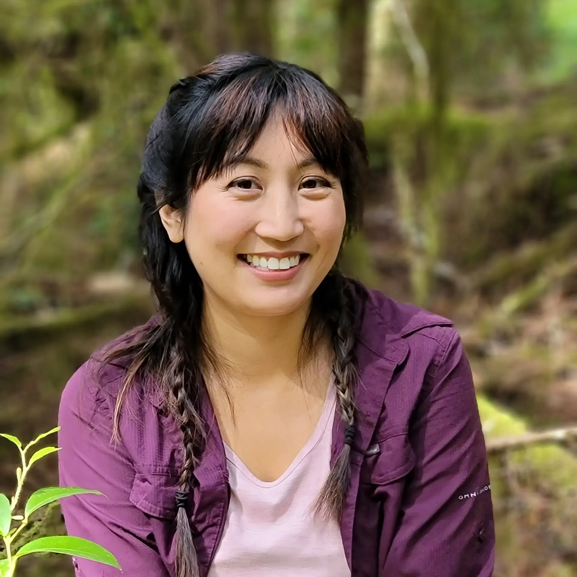 Portrait of Alannah Cross sitting outdoors in a forest setting, wearing a purple jacket over a light-colored shirt, with green foliage and trees softly blurred in the background.
