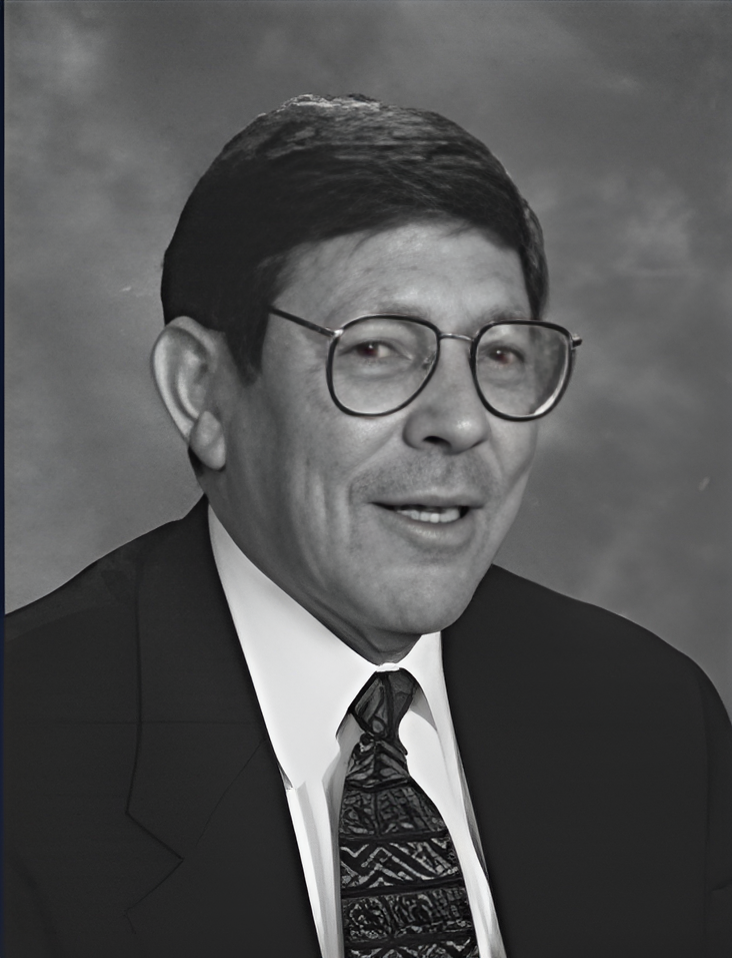 Black-and-white studio portrait of Dr. Ed Command wearing glasses, a suit jacket, white shirt, and patterned tie, facing slightly to the side against a neutral backdrop