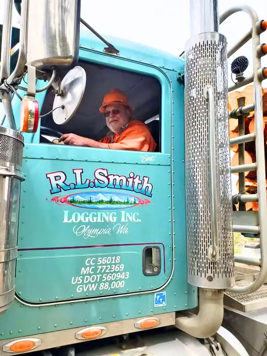A person seated inside a teal R.L. Smith Logging Inc. truck, shown from the side. The truck displays company branding and regulatory information, including “R.L. Smith Logging Inc., Olympia WA,” along with vehicle details printed on the door.