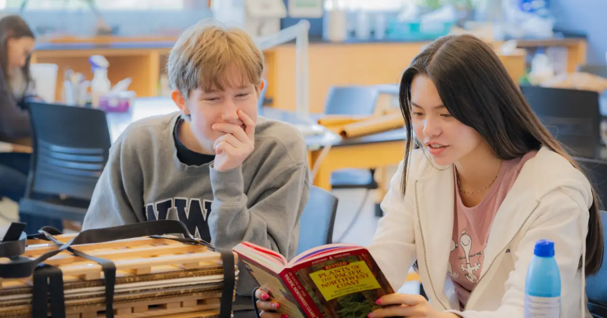 Two students sit at a table reading a book together in a classroom or study space.