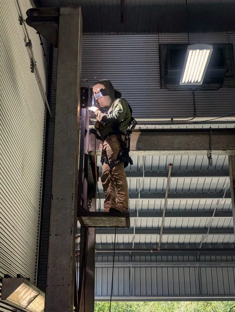 A welding student works on a steel structure inside Grays Harbor College’s welding facility. Standing on an elevated platform and secured with a safety harness, the student performs a weld on a vertical steel beam while sparks illuminate the work area. The industrial training space features exposed steel framing, high ceilings, overhead lighting, and large metal wall panels.