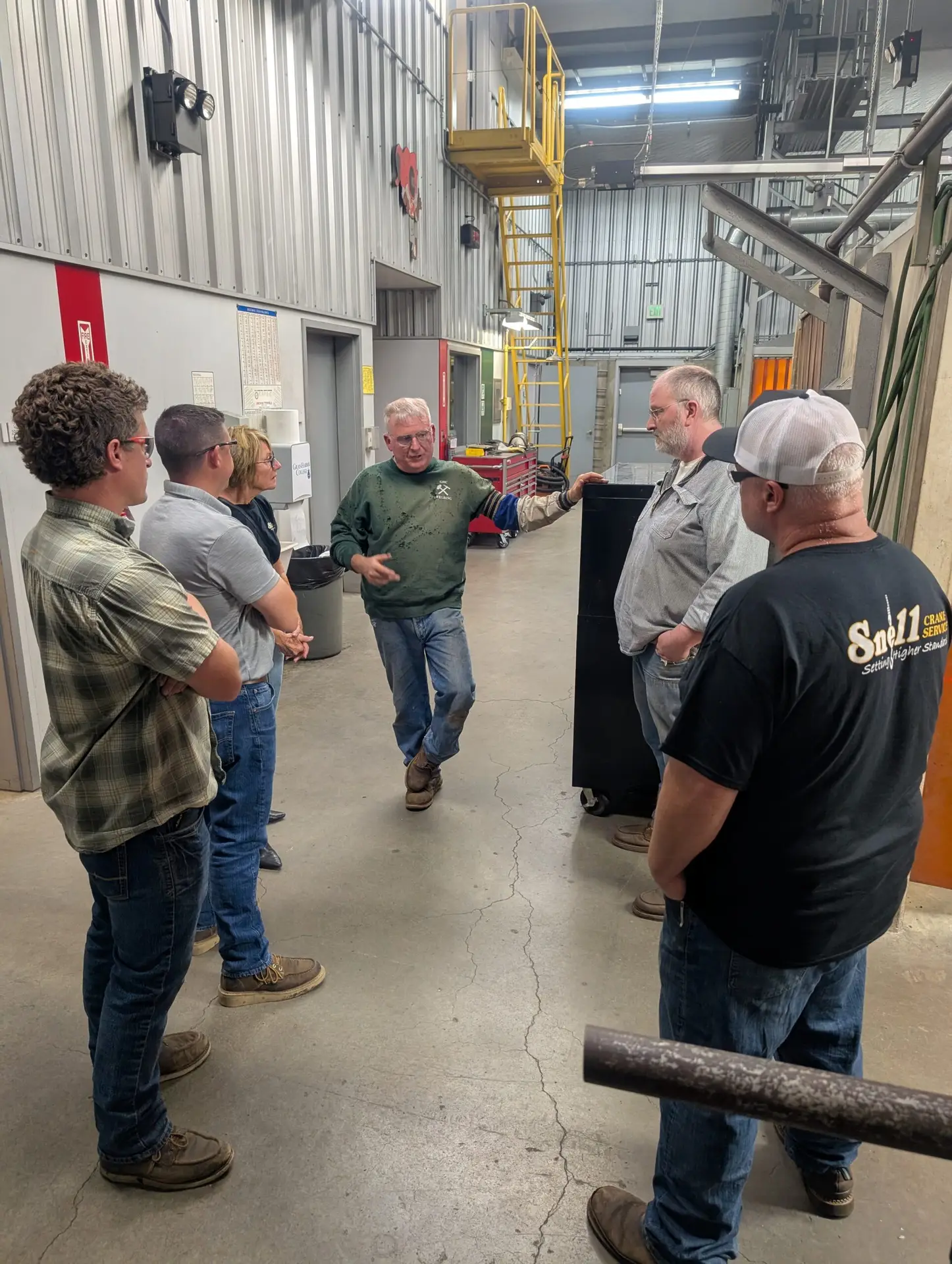 A welding instructor leads a small group tour inside Grays Harbor College’s welding facility, explaining equipment and training processes. The group stands in a spacious industrial shop with metal walls, welding stations, overhead structures, and safety equipment visible in the background while listening to the presentation