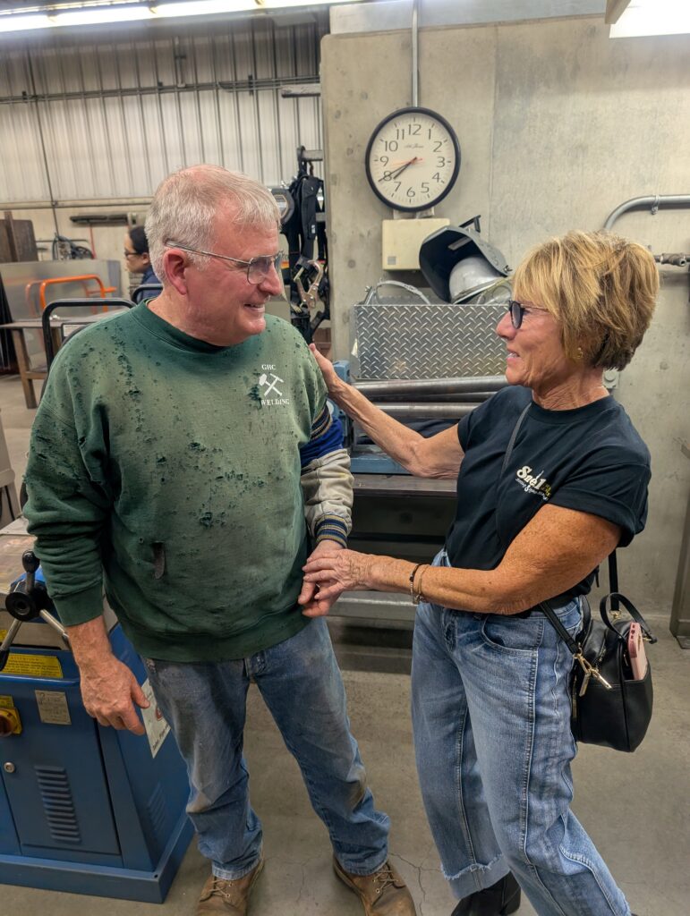 GHC welding instructor Rod McDonald visits with Carrie Snell inside Grays Harbor College's welding shop. Standing beside fabrication equipment, McDonald and Snell talk during a tour of the welding facility, with tools, machinery, and training equipment visible in the background