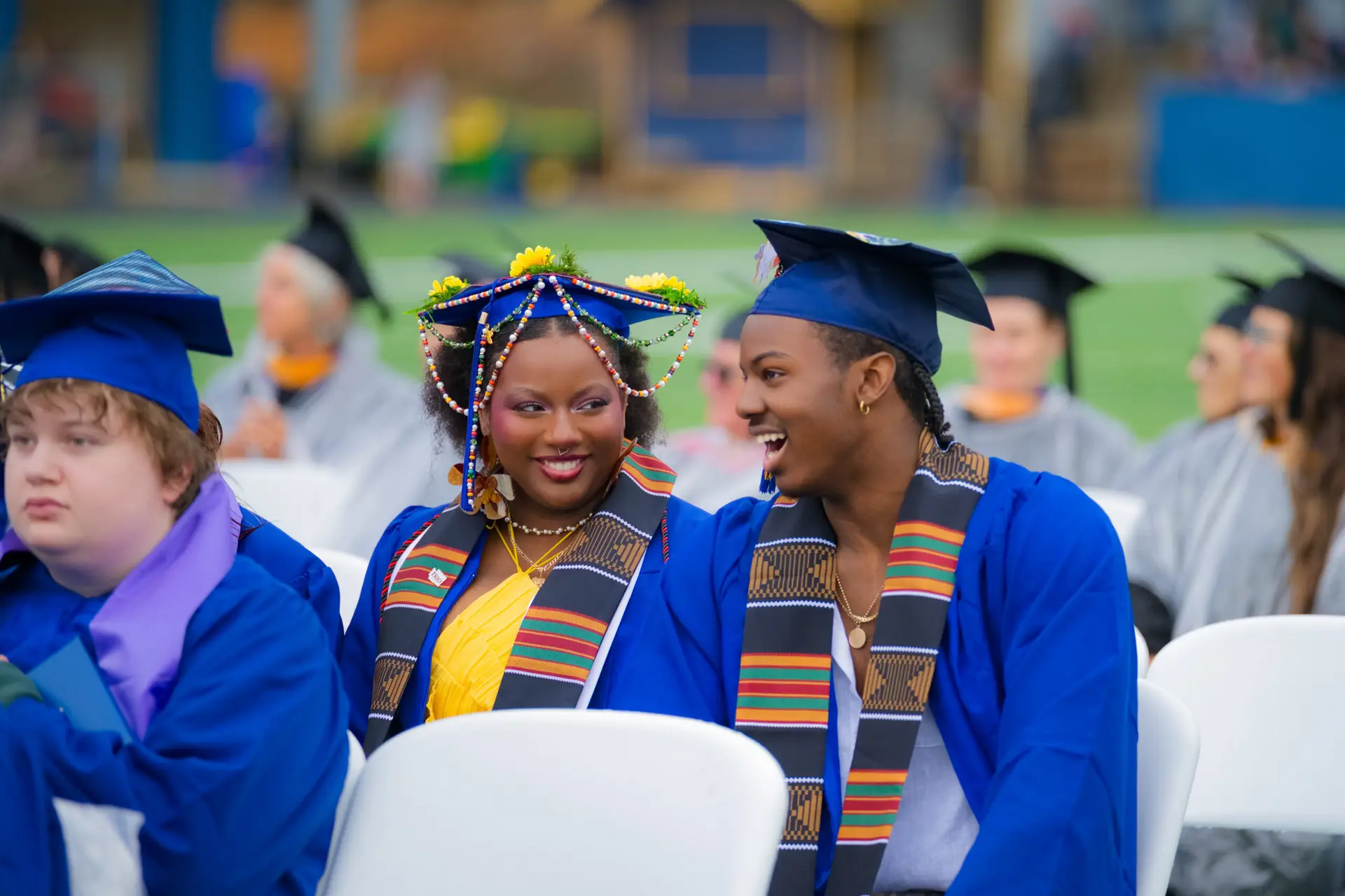 Graduates in blue caps and gowns seated at a commencement ceremony, wearing colorful stoles and decorated graduation caps